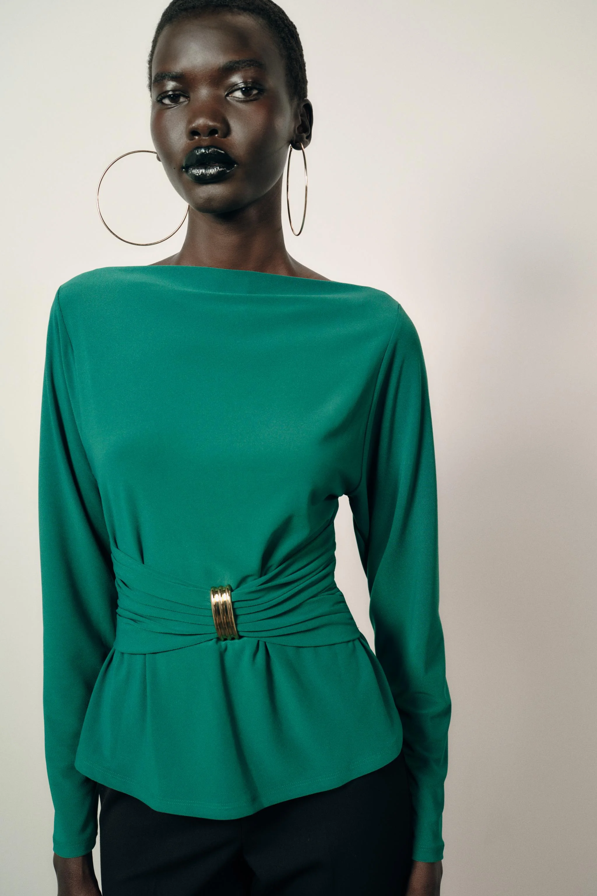 تاپ یقه قایقی با آستین بلند. دارای جزئیات چین دار در قسمت کمر با یک تکه فلزی تزئینی و لبه گرد.