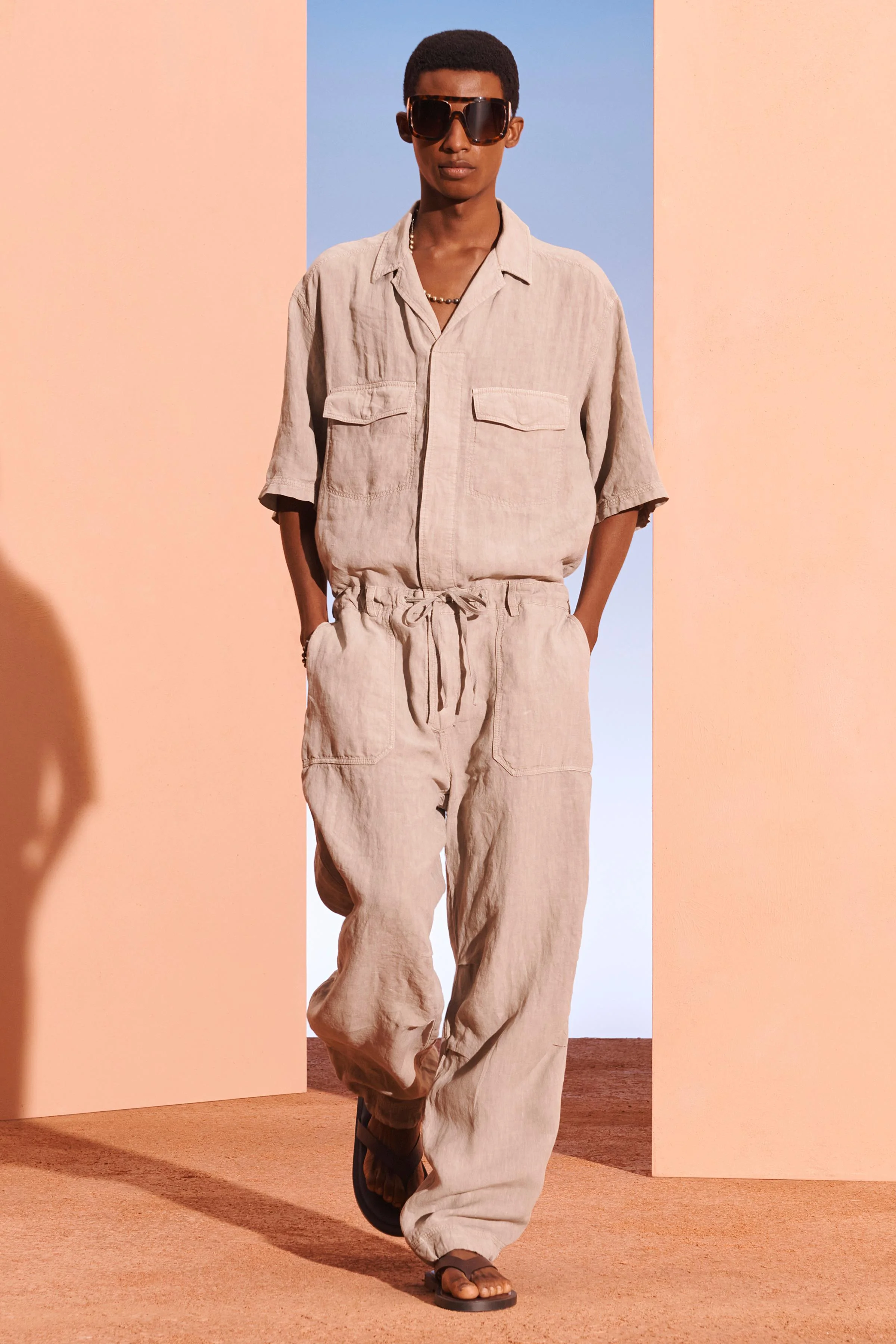 سرهمی لینن بزرگ با یقه بولینگ و آستین کوتاه. دارای جیب های وصله ای با فلپ روی سینه، کمر کشی قابل تنظیم با بند کشی، و جیب های وصله ای در جلو و پشت شلوار. دارای پلیسه در زانوها و سجاف قابل تنظیم با بند کشی در طرفین. جلو زیپ و دکمه با یک سجاف پنهان بسته می شود.